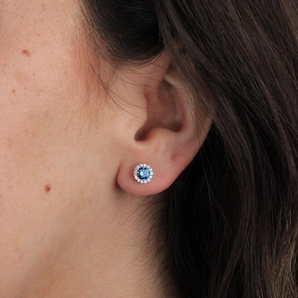 Pendientes en oro blanco de 18 quilates con un circonita azul con orla de circonitas incoloras.
